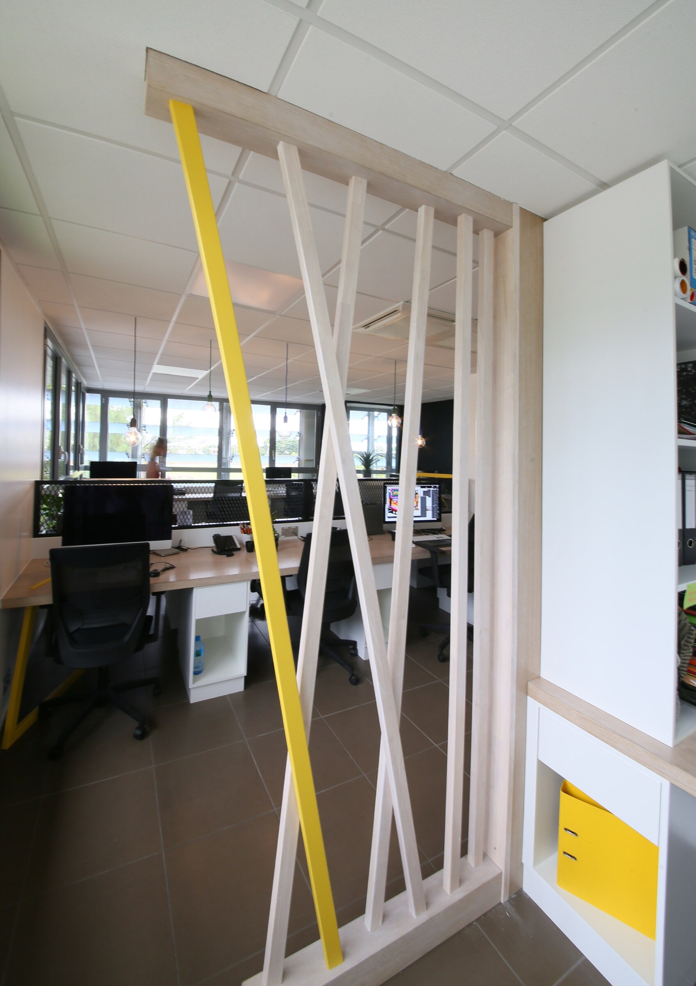 Agence de communication, bureaux — cloison à claire-voie en bois clair et latte jaune séparant l'accueil de l'open space