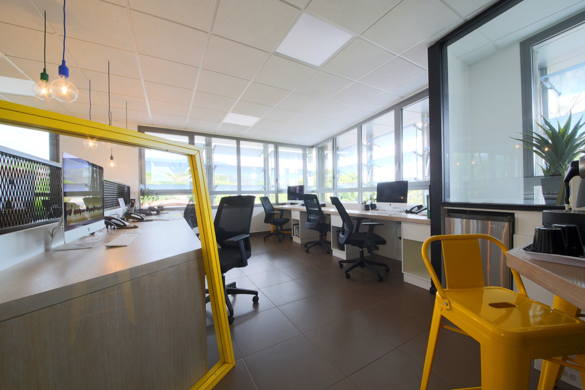 Agence de communication, bureaux — open space en longueur le long des baies vitrées, cadre jaune et tabouret jaune au premier plan