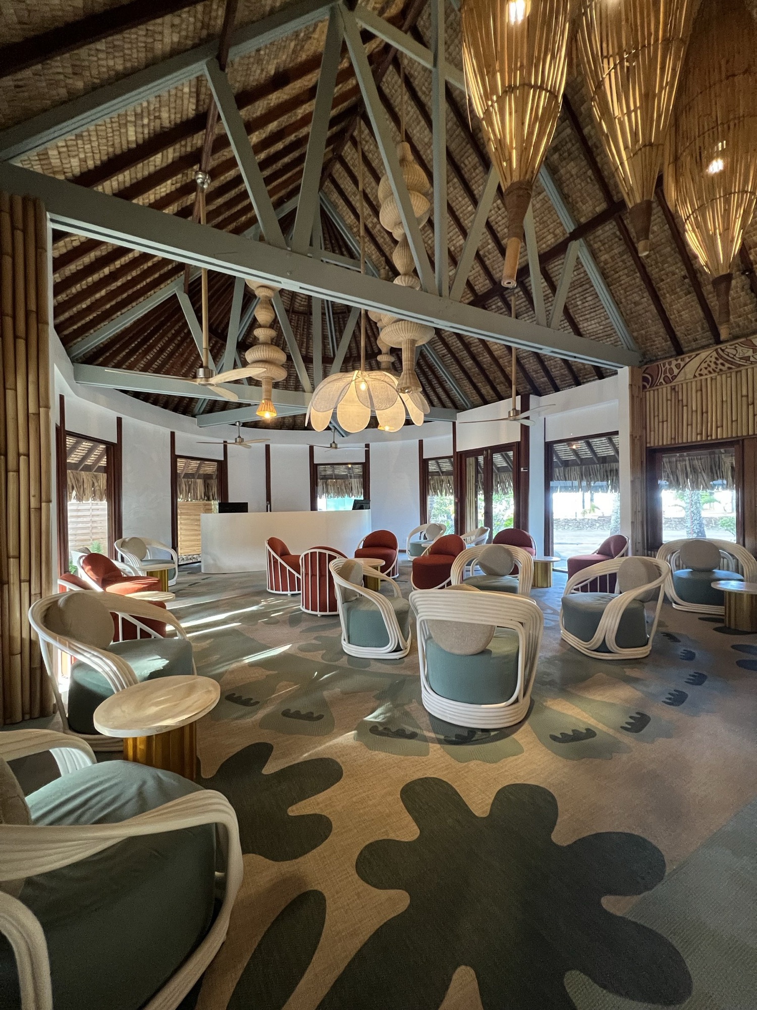 InterContinental Bora Bora, lobby — salon lounge : fauteuils en rotin blanc et velours terracotta sur un tapis à motifs végétaux, banque d'accueil au fond