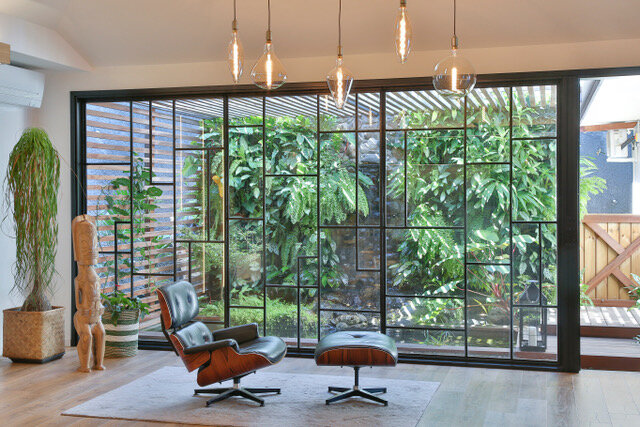 Maison particulière — salon : grande verrière en acier noir sur le jardin, fauteuil et ottoman Eames