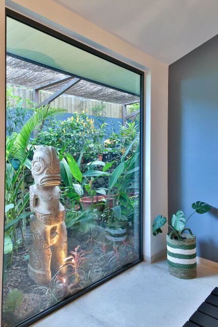 Maison particulière — baie vitrée cadrant un tiki sculpté au cœur du jardin tropical